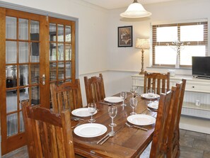 Una mesa de comedor preparada para seis personas, con sillas de madera, una araña y un televisor al fondo.