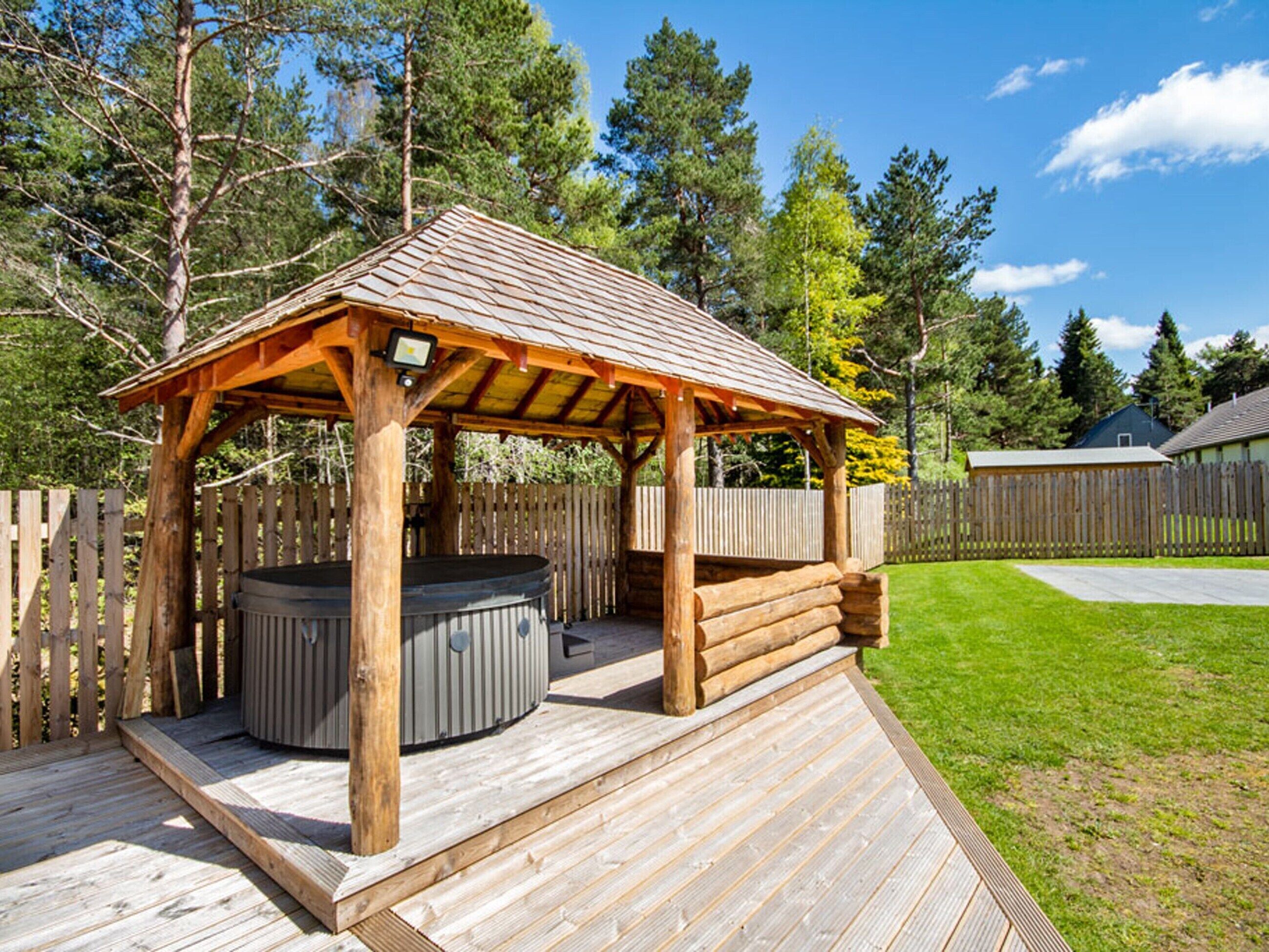Un área de hidromasaje exterior de madera con techo de pérgola, rodeada por una terraza de madera y una valla de madera.