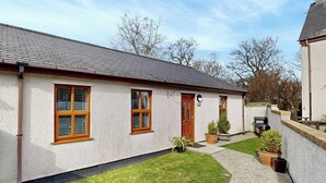 Ein eingeschossiges Haus mit weißer Fassade, einer roten Tür und einem kleinen Vorgarten mit Rasen und Blumentöpfen.