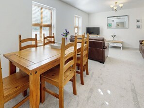Un comedor con una mesa y sillas de madera, un sofá marrón, un televisor y una pequeña mesita auxiliar blanca con un florero.