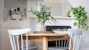 A dining area with a wooden table surrounded by white chairs, a vase with green foliage, and a white shelf with books and a power strip. - Seaside Apartment-UK2801 (Aberystwyth)