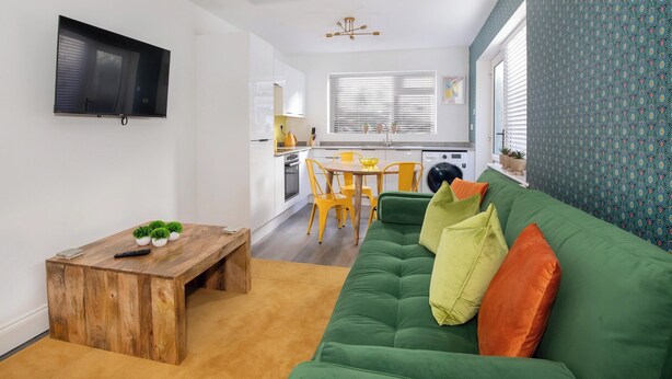 Uma sala de estar moderna com um sofá verde, mesa de centro de madeira e cadeiras amarelas. Cozinha ao fundo com armários brancos e uma janela com persianas.