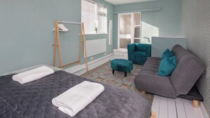 Quarto com cama, sofá cinzento, poltrona verde-água e janela com persianas.