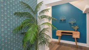 Quarto com papel de parede estampado, uma planta verde grande, uma escrivaninha de madeira e uma parede azul de destaque.