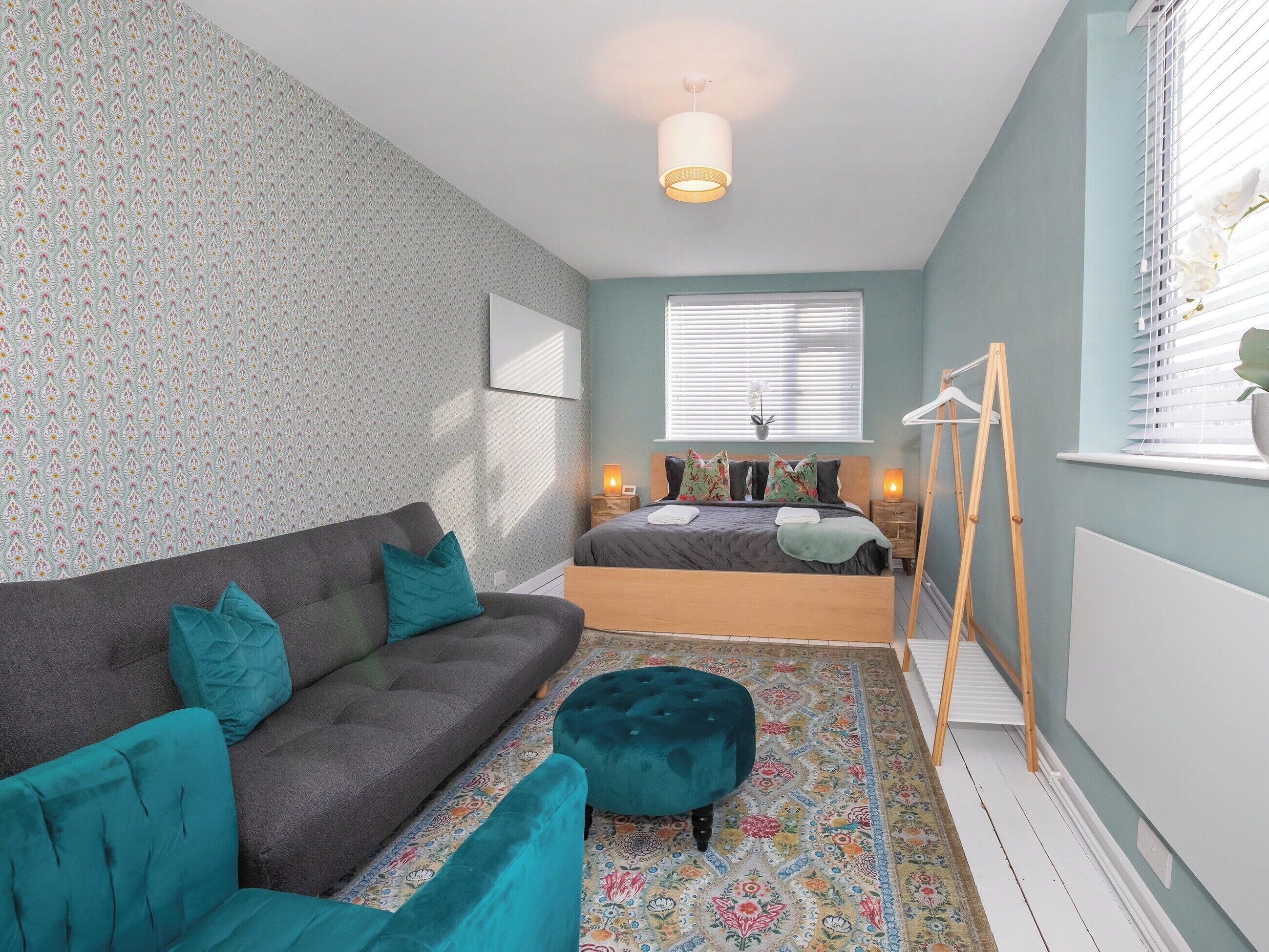 Une chambre avec un canapé gris, des coussins turquoise, un tapis à motifs, un lit avec des oreillers à fleurs, une table de chevet avec une lampe et une échelle en bois.