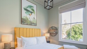 A neatly made bed with white linens, a yellow headboard, and a yellow textured throw blanket. There is a round wall shelf with two potted plants, a lamp on a bedside table, and a window with a view of greenery outside. - 3 bedroom accommodation in York (York)