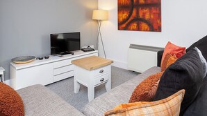 Een moderne woonkamer met een flatscreen televisie, een witte entertainmentkast, een klein houten tafeltje, een grijze bank met oranje kussens, en een groot abstract schilderij aan de muur.