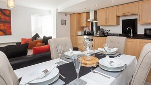 Een eetgedeelte met een tafel voor vier personen, een keuken op de achtergrond en een woonkamer met een bank en kussens.