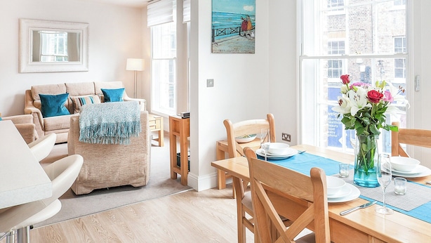 Ett ljust vardagsrum med en beige soffa, ett matbord dukat med tallrikar och en vas med blommor, samt en tavla som föreställer en strandscen.