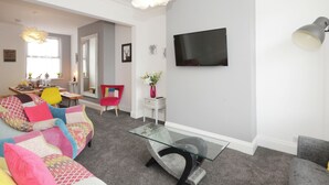 Een moderne woonkamer met een glazen salontafel, een bank met kleurrijke kussens, een eethoek met een tafel voor vier personen, en een flatscreen televisie aan de muur.
