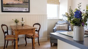 Uma área de refeições com mesa e cadeiras de madeira, um sofá com almofadas, uma janela com persiana estampada e um quadro na parede.