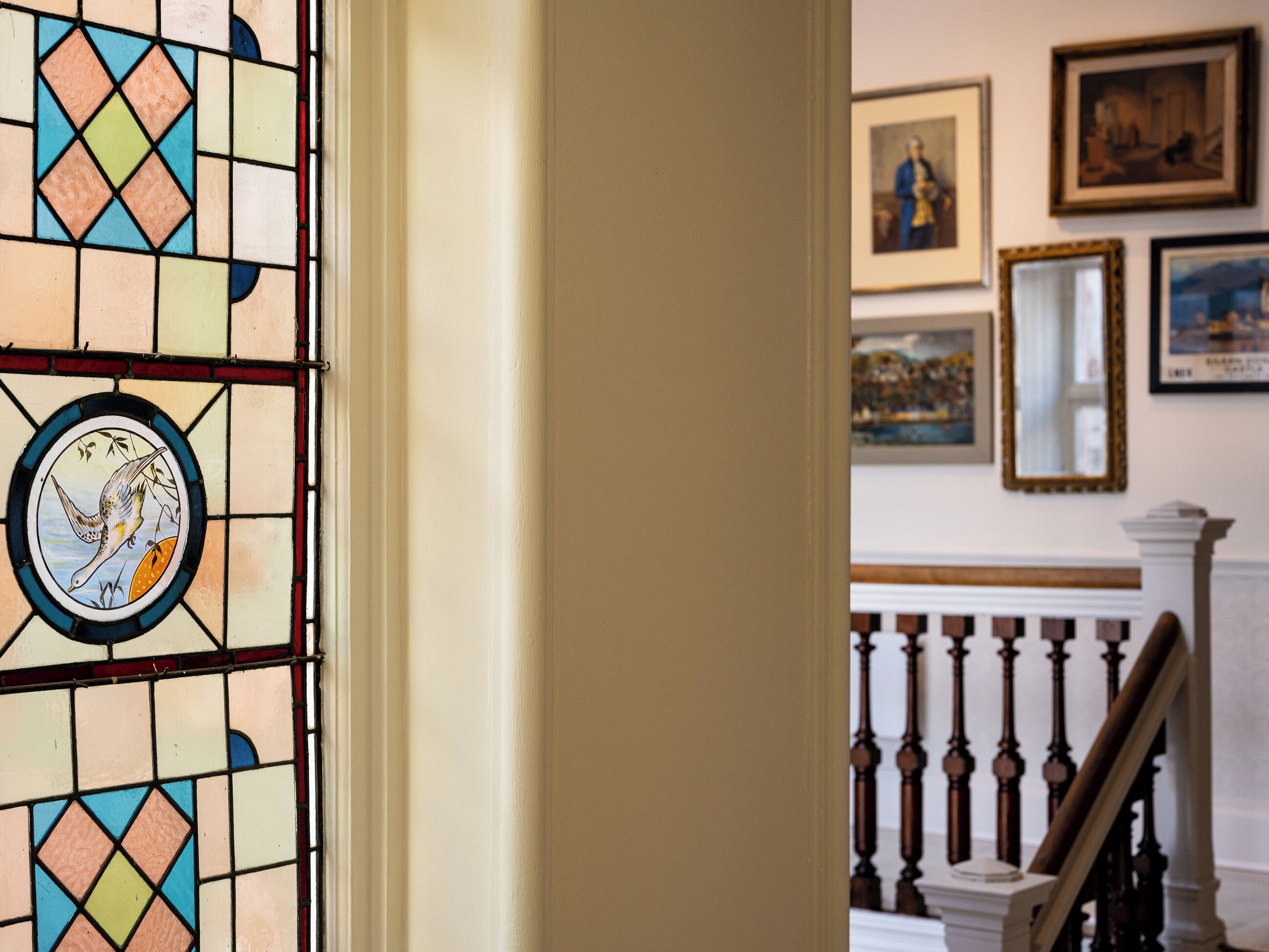 A stained glass window with a bird design, a staircase with a balustrade, and framed artwork on the wall.