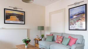 A living room with a grey sofa, a wooden coffee table, a framed landscape painting, a lamp, and a vase with flowers. - 2 bedroom accommodation in Inverness (Inverness)