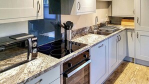 A modern kitchen with white cabinets, a black induction cooktop, a stainless steel sink, and a wooden floor. - Oakbank Cottage - UK7574 (Pitlochry)