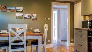 A dining area with a wooden table and white chairs, a kitchen with a microwave, and a door leading to another room. - Oakbank Cottage - UK7574 (Pitlochry)