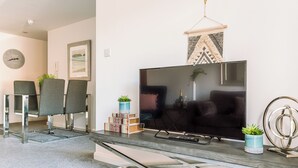 Una sala de estar moderna con un televisor de pantalla plana, dos sillones grises, una mesita con una planta y un adorno colgante en la pared.