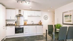 Una cocina moderna con armarios blancos, una campana de acero inoxidable, horno empotrado, un lavamanos y una zona de comedor con mesa y sillas de vidrio.