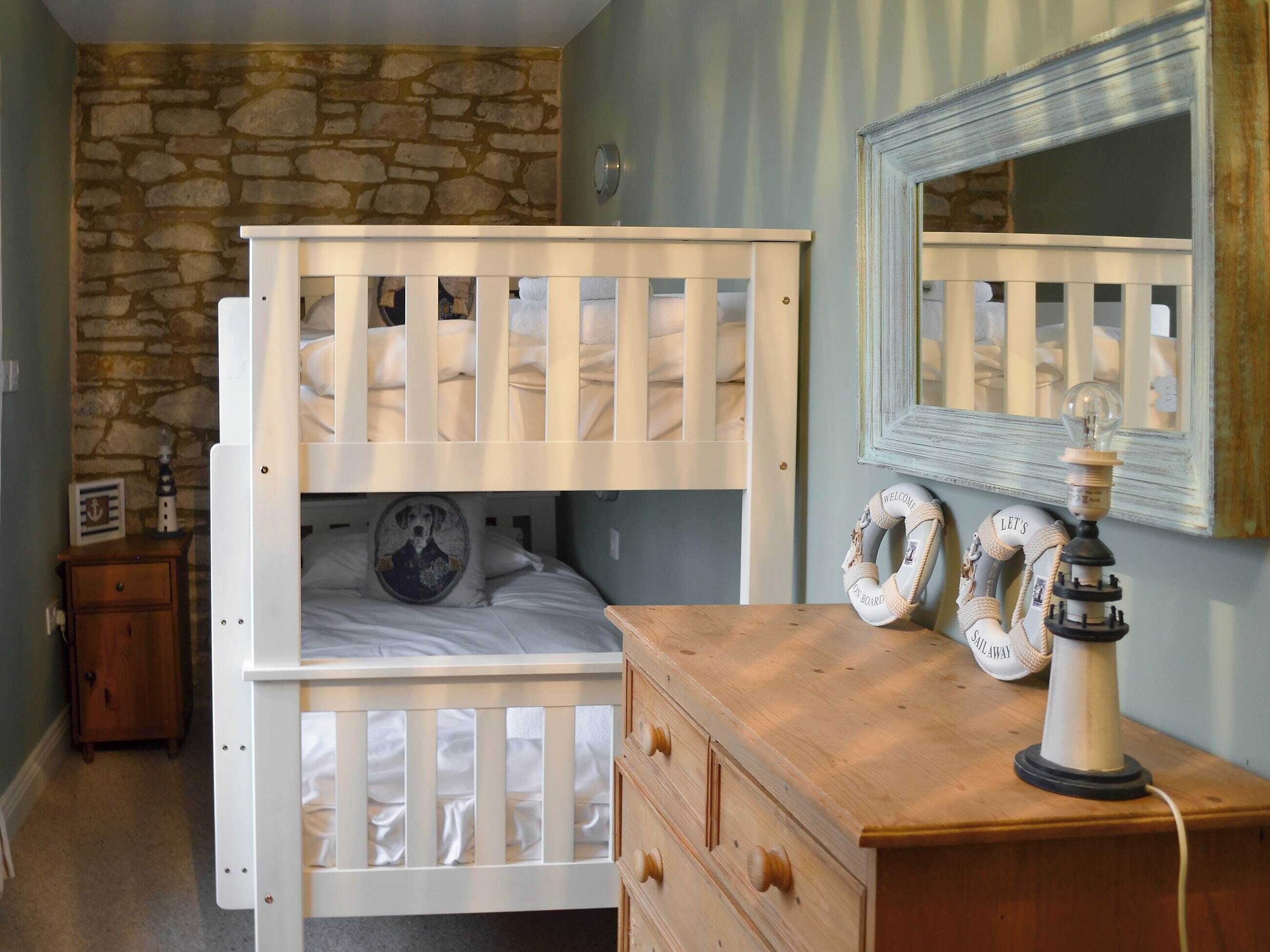 Un lit superposé avec une structure en bois et un lit inférieur agrémenté d’un coussin orné d’un motif canin. Une commode en bois avec des tiroirs se trouve à côté du lit, et une lampe avec une ampoule est posée sur la commode. Un mur en pierre et un miroir se trouvent à l’arrière-plan.