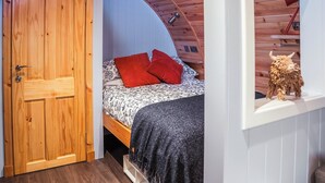 Un dormitorio con puerta de madera, cama con ropa de cama estampada y almohadas rojas, una manta de color negro y techo de madera.