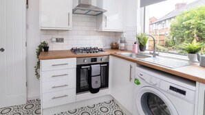Une cuisine compacte avec des meubles blancs, une cuisinière à gaz, un four, un lave-linge et une fenêtre donnant sur un jardin.