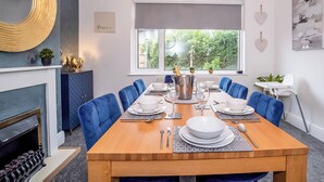 Une salle à manger avec une table en bois dressée pour six personnes, des chaises à l’assise bleue, une cheminée et une vue sur la verdure à l’extérieur.