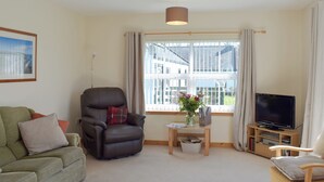 A living room with a green sofa, a leather armchair, a wooden coffee table with a vase of flowers, a television on a wooden stand, and a window with curtains. - Willow Cottage - UK30684 (Sandhead)