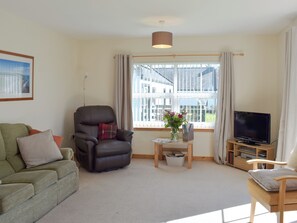 Ein Wohnzimmer mit einem grünen Sofa, einem Ledersessel, einem Couchtisch aus Holz mit einem Blumenstrauß, einem Fernseher auf einem Holzboden und einem Fenster mit Vorhängen.