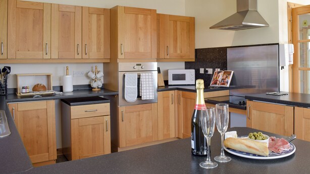 A modern kitchen with wooden cabinets, stainless steel appliances, and a central island with a bottle of champagne, glasses, and a plate of food. - Willow Cottage - UK30684 (Sandhead)
