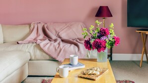 En hyggelig stue med en beige sofa, et pink plaid, et træsofabord med en kop, småkager og en vase med blomster.