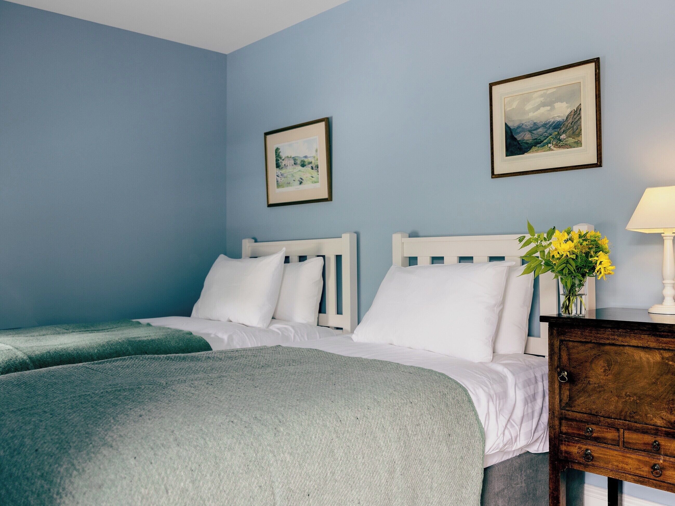 A bedroom with two beds, a nightstand, a lamp, and two framed pictures on the wall.