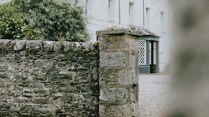 A stone wall with a small gate, a white building with multiple windows, and a green bush in the foreground.