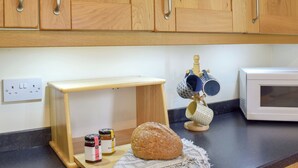 Une cuisine avec des placards en bois, une petite table en bois, des pots de confiture, une miche de pain, une cafetière et un four à micro-ondes.