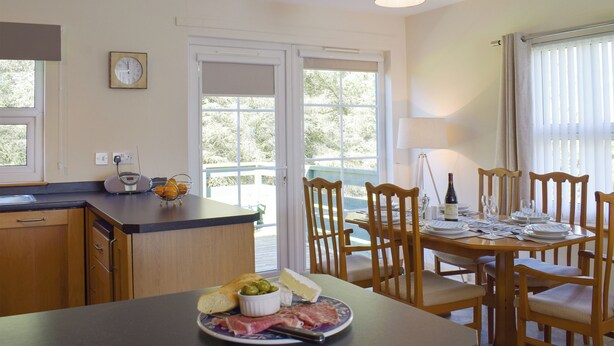 Un coin repas avec une table dressée pour deux personnes, un comptoir de cuisine avec une balance et des oranges, et une fenêtre donnant sur un paysage verdoyant.