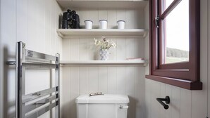 Ett litet badrum med en toalett, en hylla med muggar och en vas med blommor, ett fönster och en handdukshängare.