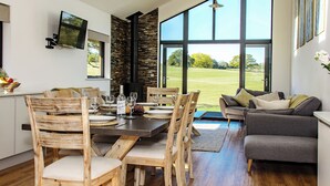 Een eetgedeelte met een houten tafel en stoelen, een bank en een groot raam met uitzicht op een groen landschap.