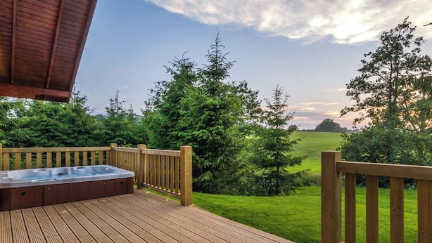 A wooden deck with a hot tub, surrounded by greenery and a view of a field. - Rawlsbury Lodge - UK31007 (Ansty, near Dorchester)