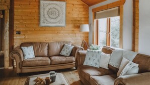 A cozy room with wooden walls, a brown leather sofa, a wooden coffee table with a tray of pastries, and a large window with a view of trees. - Rawlsbury Lodge - UK31007 (Ansty, near Dorchester)