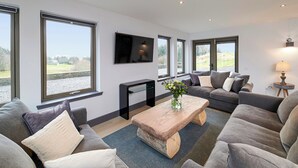 Una sala de estar moderna con ventanas amplias, un televisor de pantalla plana, una mesa de centro de madera y sofás grises.