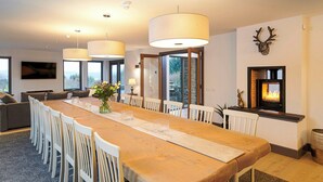 Un comedor moderno con una mesa de madera larga, sillas blancas y lámparas colgantes. Hay una chimenea con un adorno de cabeza de ciervo en la pared.