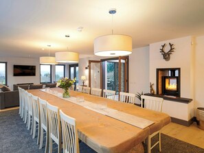A modern dining room with a long wooden table, white chairs, and pendant lights. There is a fireplace with a deer head decoration on the wall. - 5 bedroom accommodation in Drumnadrochit (Drumnadrochit)