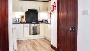 A compact kitchen with white cabinets, a black backsplash, and a wooden floor. There's a stove, microwave, and toaster on the counter. A fire extinguisher is mounted on the wall. - Merrick Cottage - UK7783 (Newton Stewart)