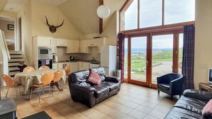 A spacious living area with a large window, a dining table with chairs, a leather sofa, and a kitchen in the background. - 3 bedroom accommodation in Lumphanan, near Banchory (Lumphanan, near Banchory)