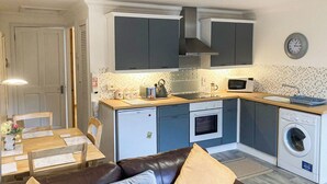 Una cocina compacta con refrigerador, horno, microondas y lavadora. Hay una mesa de comedor con sillas y un sofá de cuero marrón con cojines.
