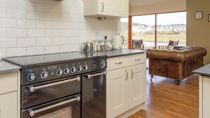 Uma cozinha moderna com eletrodomésticos de aço inoxidável, armários brancos e fogão de vitrocerâmica. É possível ver um lago e algumas casas através de grandes janelas.