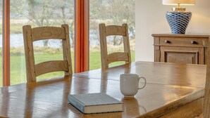 Uma mesa de madeira com duas cadeiras, uma caneca branca e um livro. Uma luminária e um vaso estão em um armário ao lado da mesa.
