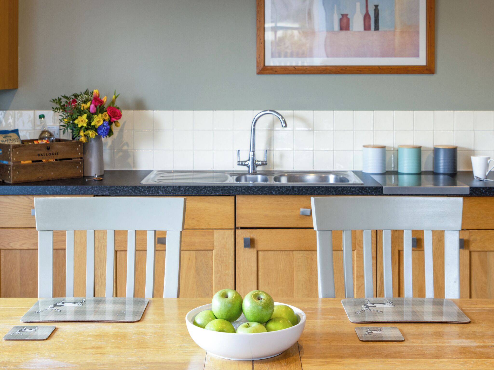A kitchen with a sink, two chairs, a table with a bowl of green apples, a vase with flowers, and a framed picture on the wall.