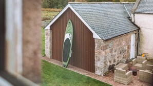 Un edificio de piedra con una tabla de surf verde en la pared lateral, una valla de madera y un patio cubierto de césped.