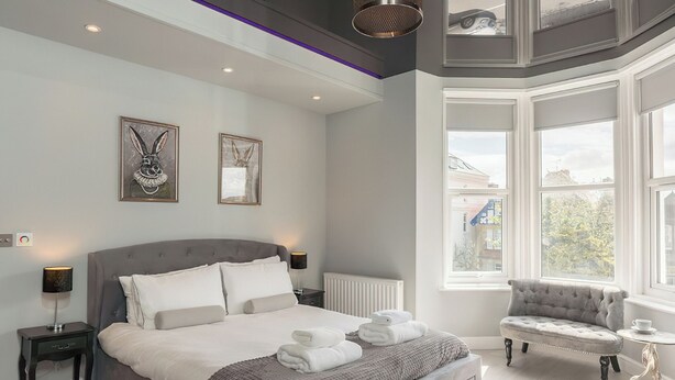 A modern bedroom with a large bed, bedside tables, a chair, and a window with a view. - Coastal Gem - UK2773 (Llandudno)
