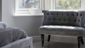 A grey tufted armchair with wooden legs, positioned near a window with a view of buildings and trees. - Coastal Gem - UK2773 (Llandudno)