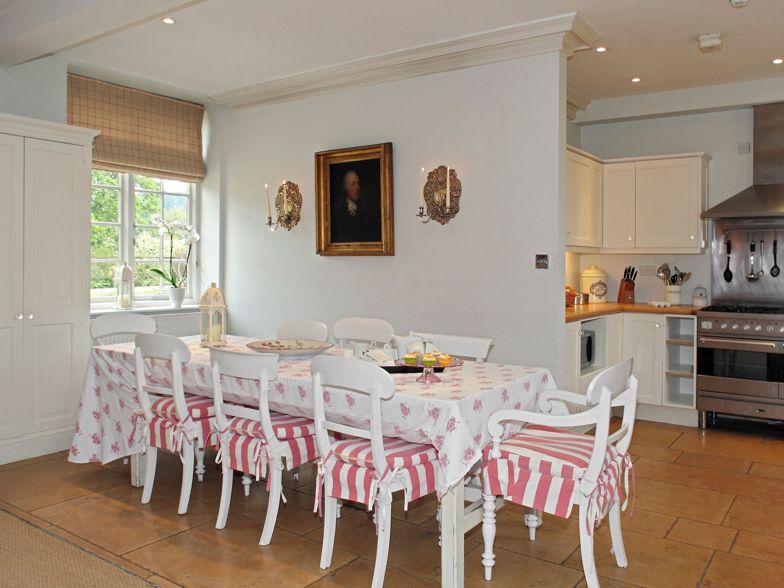 Comedor con una mesa dispuesta para seis comensales, sillas blancas con cubresal rosa y blanco a rayas, un cuadro sobre la mesa, y una zona de cocina con electrodomésticos modernos.